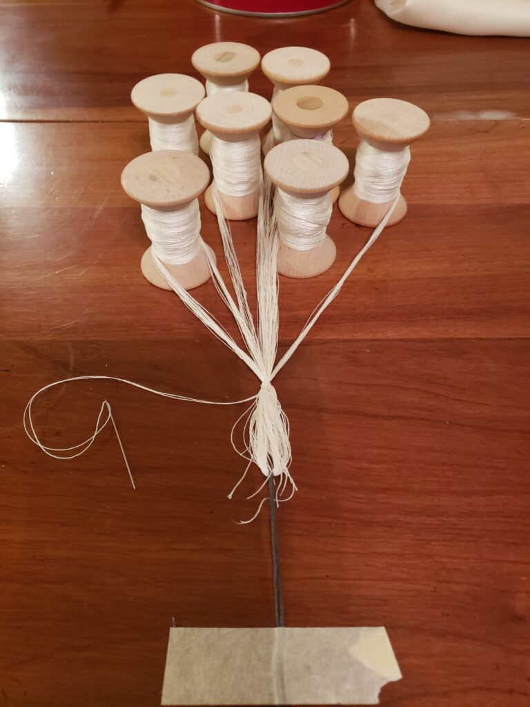 8 wooden spools with white strands made up of multiple threads THe strands are gathered together and bound. Needle and threadextend to the left of the bound spot. The strands have looped ends which have a dark colored string tying them together in a lark's head knot. The dark string is taped to the wooden surface with a piece of masking tape.