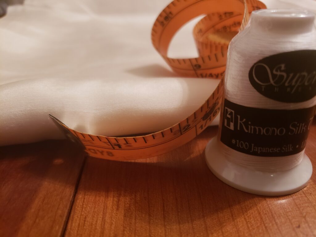 A yellow soft measuring tape curls over the fold of white silk fabric. In front of the fabric is a spool of white thread. The label reads, "Superior Thread, Kimono Silk, #100 Silk Thread"