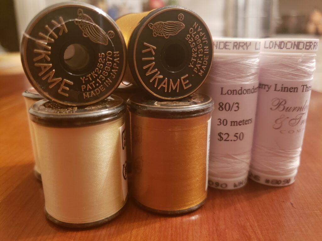 A collection of spools of thread. Left is Kinkame brand pale yellow, center is kinkame brand in gold. The kinkame spools have one spool standing on end and another laying on top with the end toward the camera. On the right are 2 spools of 80/3 Londonderry Linen thread in white from Burnley and Trowbridge .