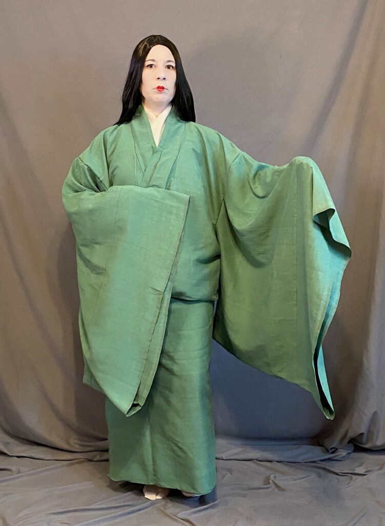 A woman in Heian make up (white face and small red lips) wearing a hitoe, belted up at the waist, with one arm outstretched to display the large sleeves.