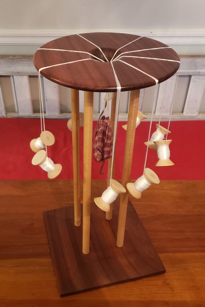 A Marudai, or Japanese braiding stand, prepped for weaving. It is a wooden device with disk top and square bottom. The top is held 2 feet above the bottom by 4 wooden dowel legs. A bag of weight hangs from the cord in the center. 8 tama hang over the edge of the mirror/round top of the marudai.