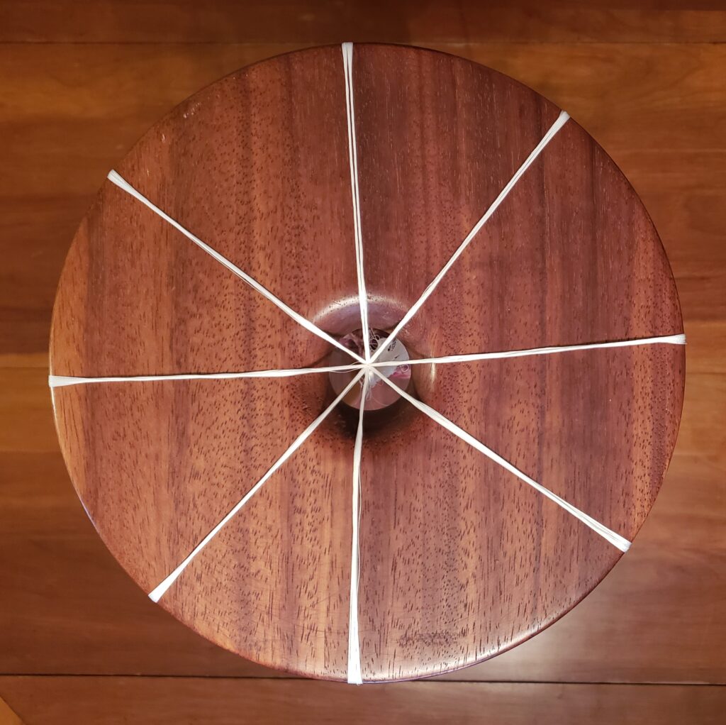 This is a top down view of a marudai set with 8 strands. we see a round wooden top divided evenly by 8 white strands that join in the center over a hole in the round top.