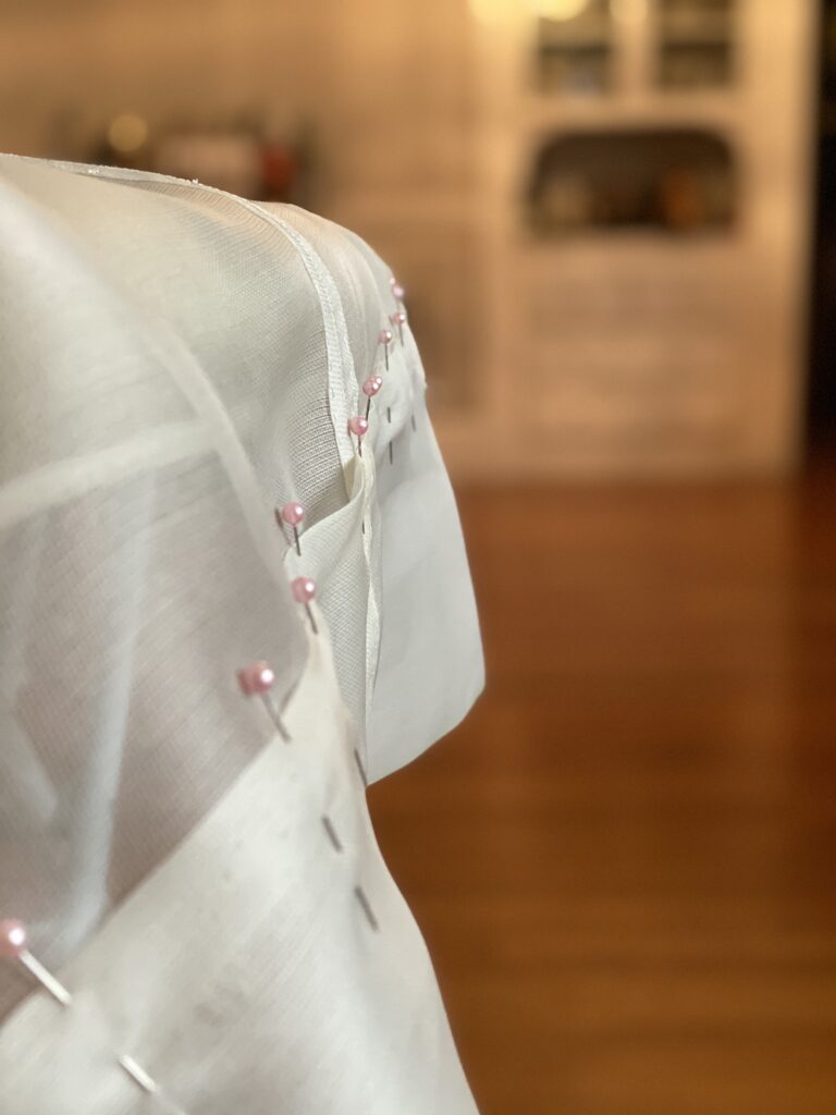 Fabric hangs over the edge of an ironing board. It has been folded back on itself and held in place by pink-headed straight pins.