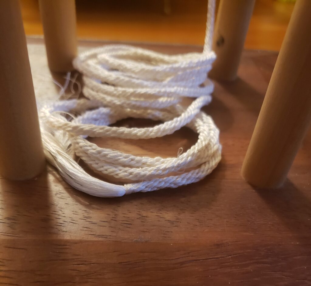 On a wooden base of a marudai rests white braided cord, coiled in tight loops between the 4 upright wooden dowel legs of the marudai.