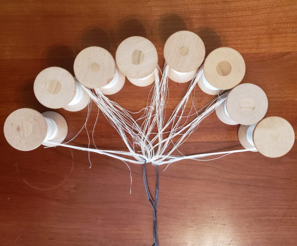 8 wooden spools with white strands made up of multiple loose threads are arranged in a rough semi-circle The looped ends of the strands extend to the center point of the semicircle and a dark grey string has been passed through the looped ends.