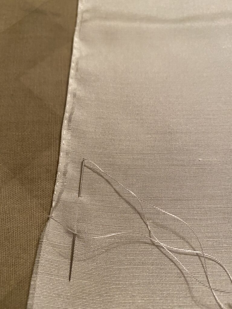 The edge of the white silk fabric has been folded over 1/16 inch and secured with a running stitch. A needle threaded with white thread is woven through the fabric.
