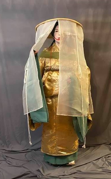 a woman in Heian make up (white face with small red lips) wearing a travelling outfit of several robes and a large, veiled hat with decorative cords