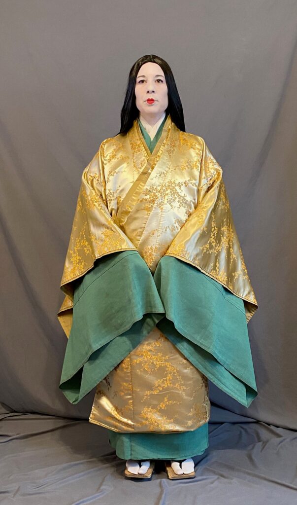 A woman in Heian make up (white face with small red lips) wearing a brocade uwagi over kosode and hitoe The longer green hitoe sleeves show vividly against the gold brocade of the uwagi